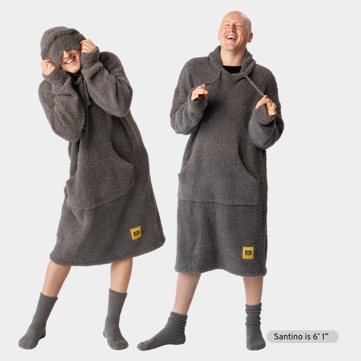 Two people wearing charcoal gray oversized hoodie blankets with drawstring hoods, styled with matching socks