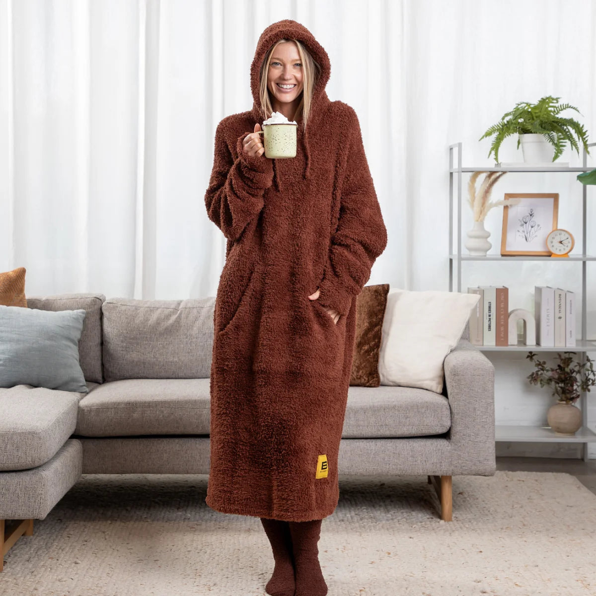 Woman wearing chocolate brown oversized blanket hoodie with hood and front pocket, holding mug in living room setting