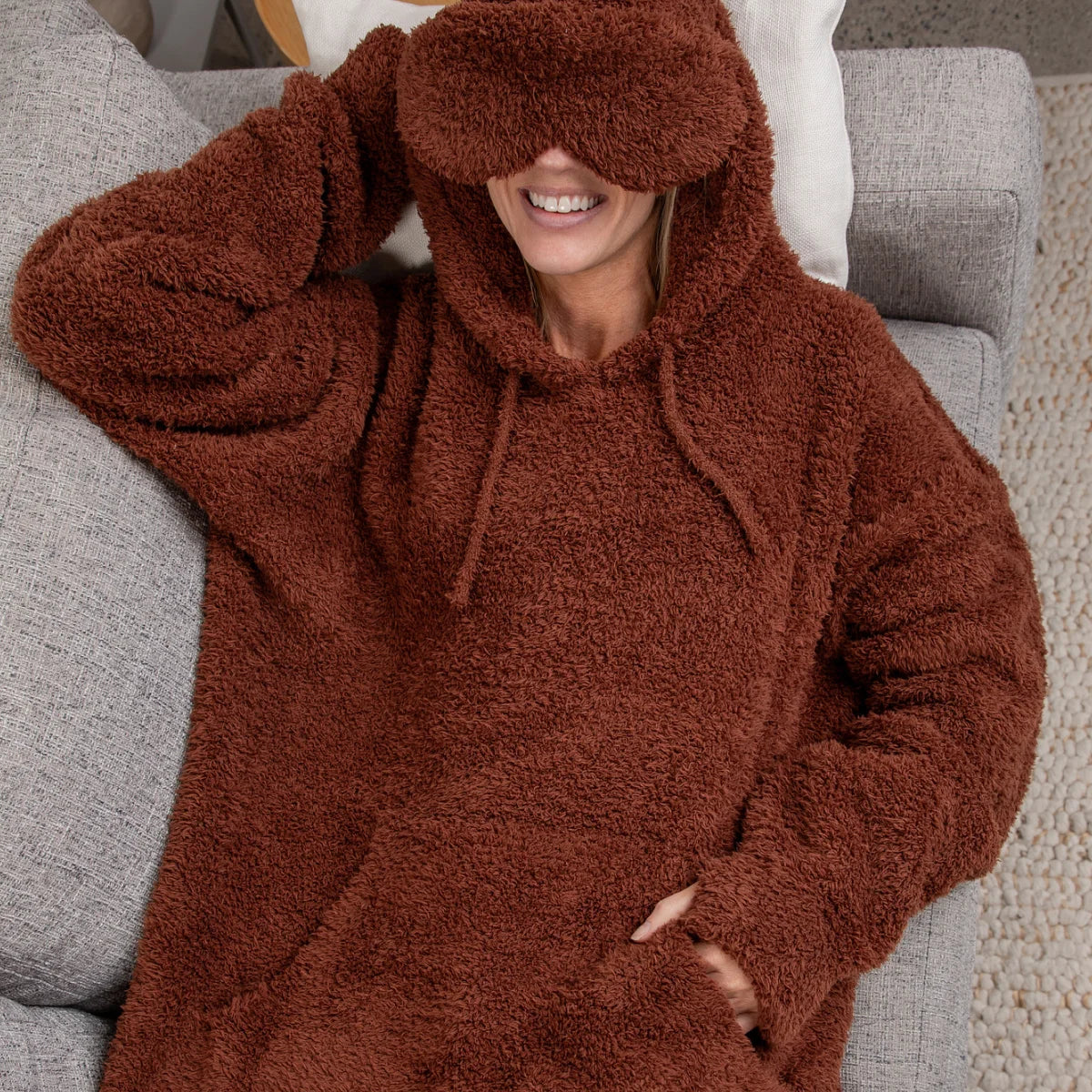 Woman wearing chocolate brown oversized plush hoodie with hood pulled over head, relaxing on gray sofa