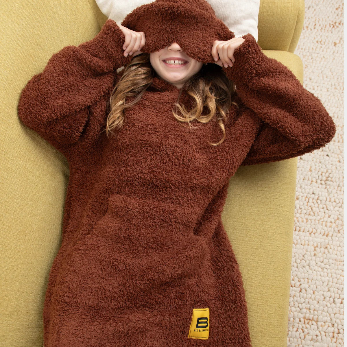 Woman wearing chocolate brown oversized fuzzy hoodie blanket draped over head on gray couch