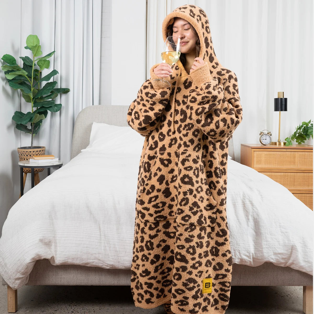Woman wearing leopard print oversized hoodie blanket on bed, sipping beverage