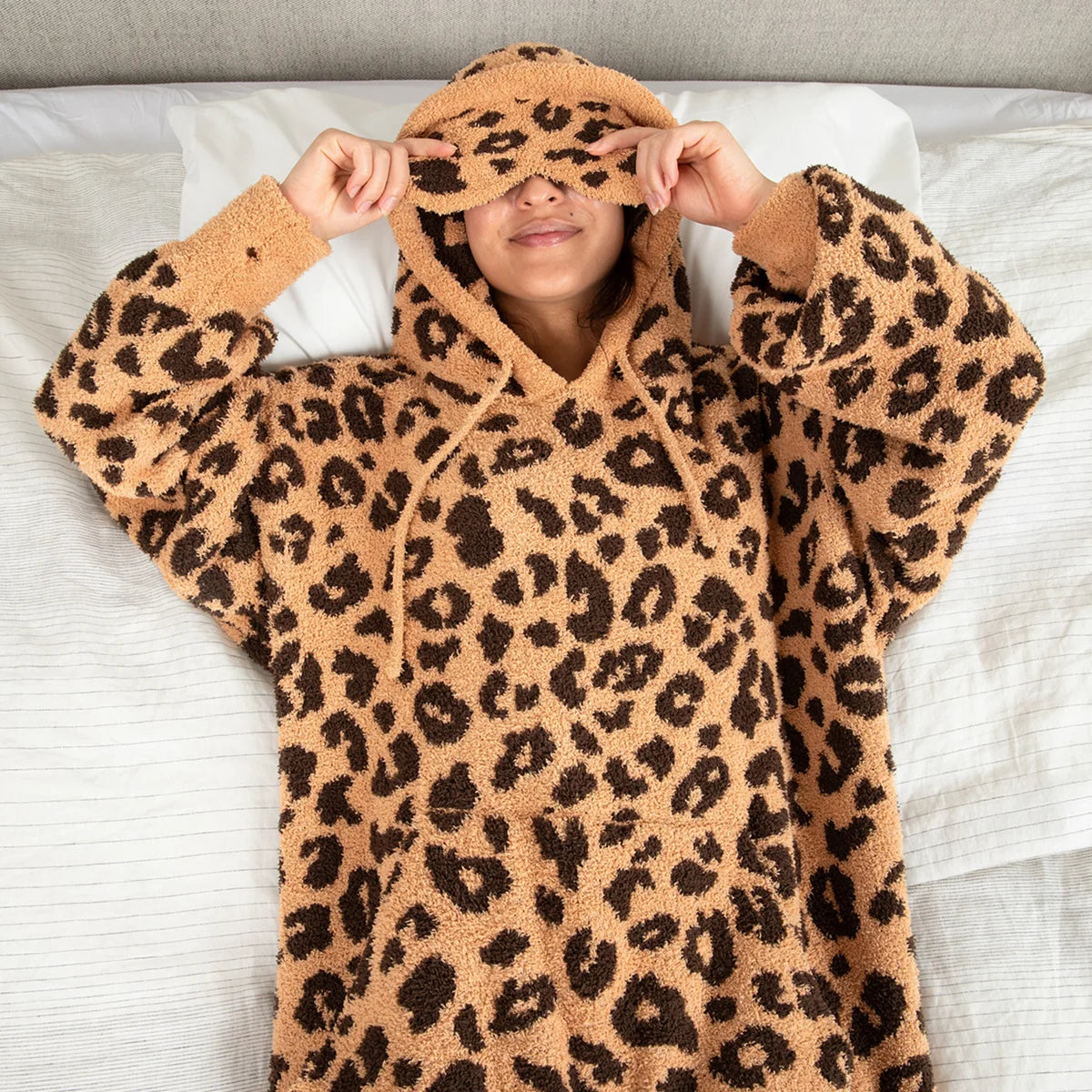 Person wearing tan leopard print oversized hoodie blanket with matching eye mask, lying on bed with white pillows