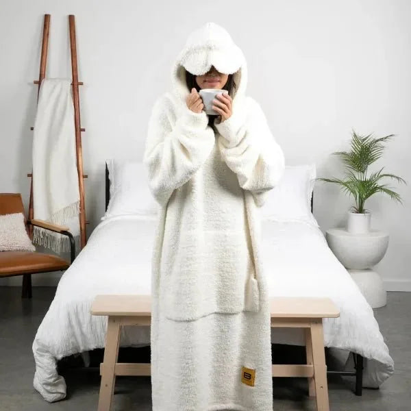 Person wearing cream oversized hoodie blanket with hood up, holding warm beverage on bed with white bedding