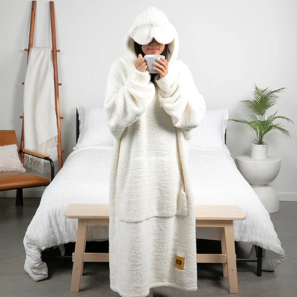Person wrapped in oversized cream hoodie blanket sitting on bed, holding mug with hood covering face