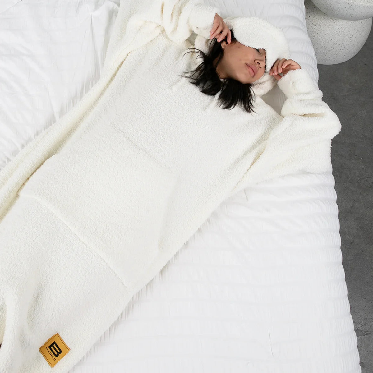 Person wearing cream hoodie blanket, cocooned in oversized cream blanket on bed, overhead view