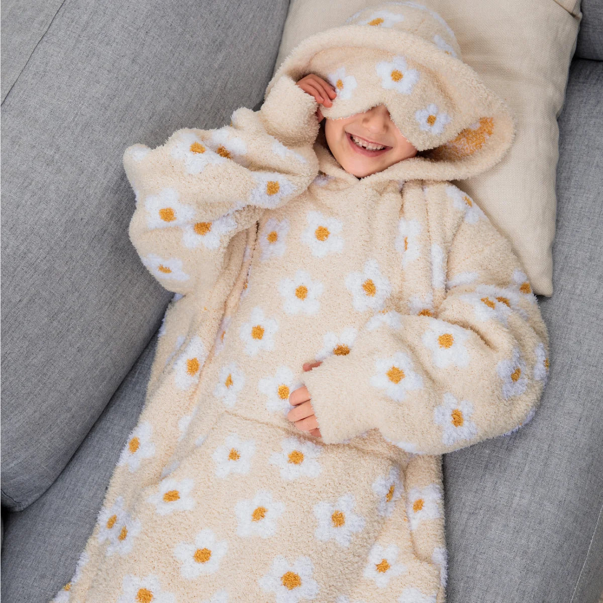 Woman wearing cream oversized blanket hoodie with yellow daisy pattern and hood, holding mug on bed