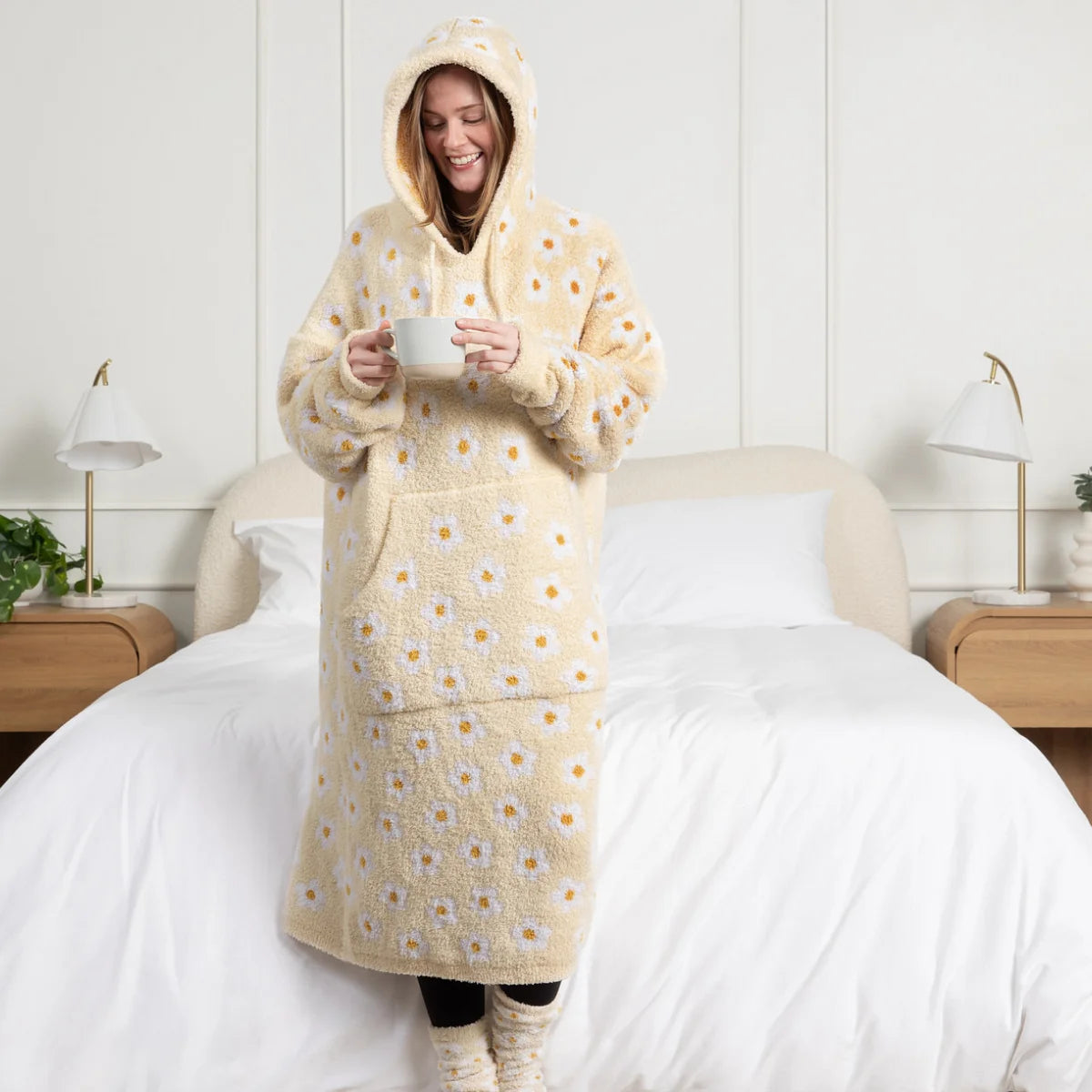 Woman wearing cream oversized hoodie blanket with yellow daisy print, holding mug on bed