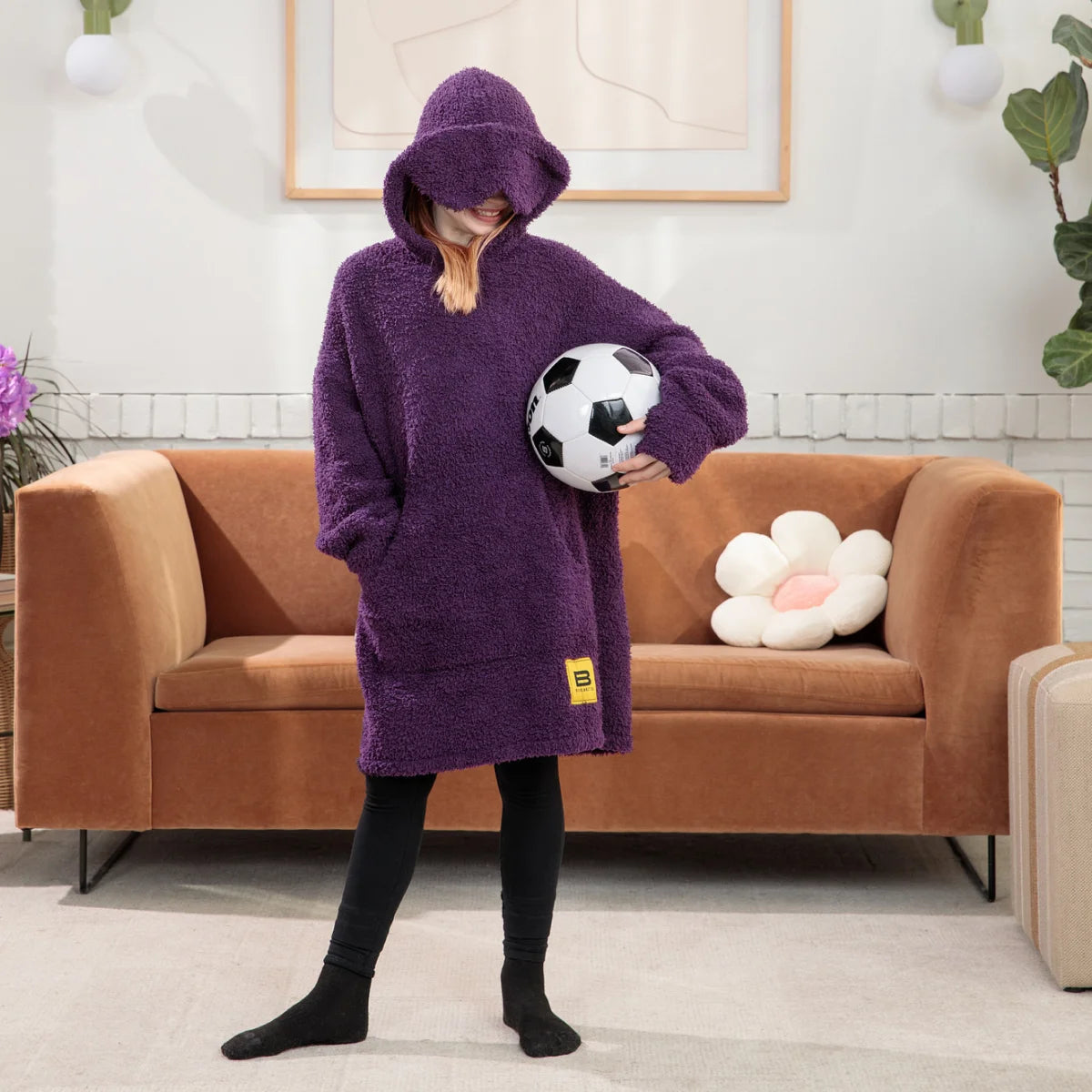 Person wearing deep purple oversized hoodie blanket with kangaroo pocket and drawstring hood while standing on bed