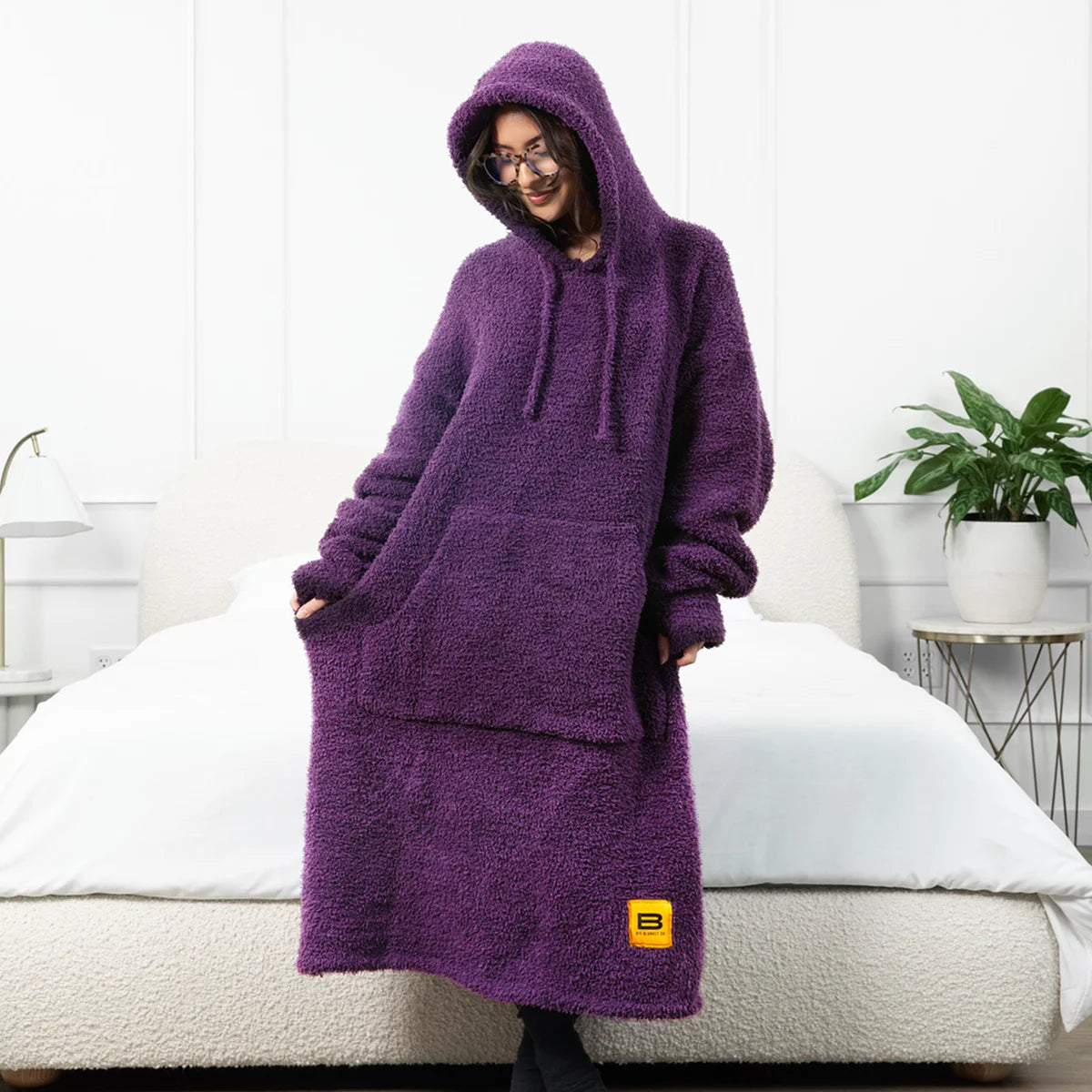 Person wearing oversized purple hoodie blanket with hood up, sitting on bed with white pillows