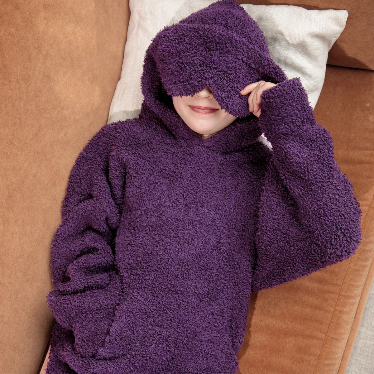 Child wrapped in deep purple oversized hoodie blanket on white bedding, pulling hood over face playfully