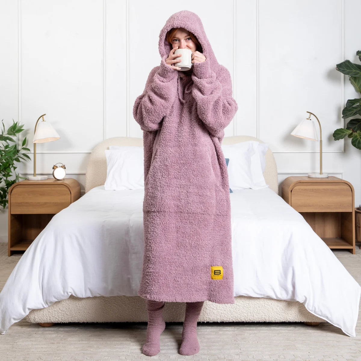 Woman wrapped in oversized purple-pink fuzzy hoodie blanket, holding mug, standing in front of bed
