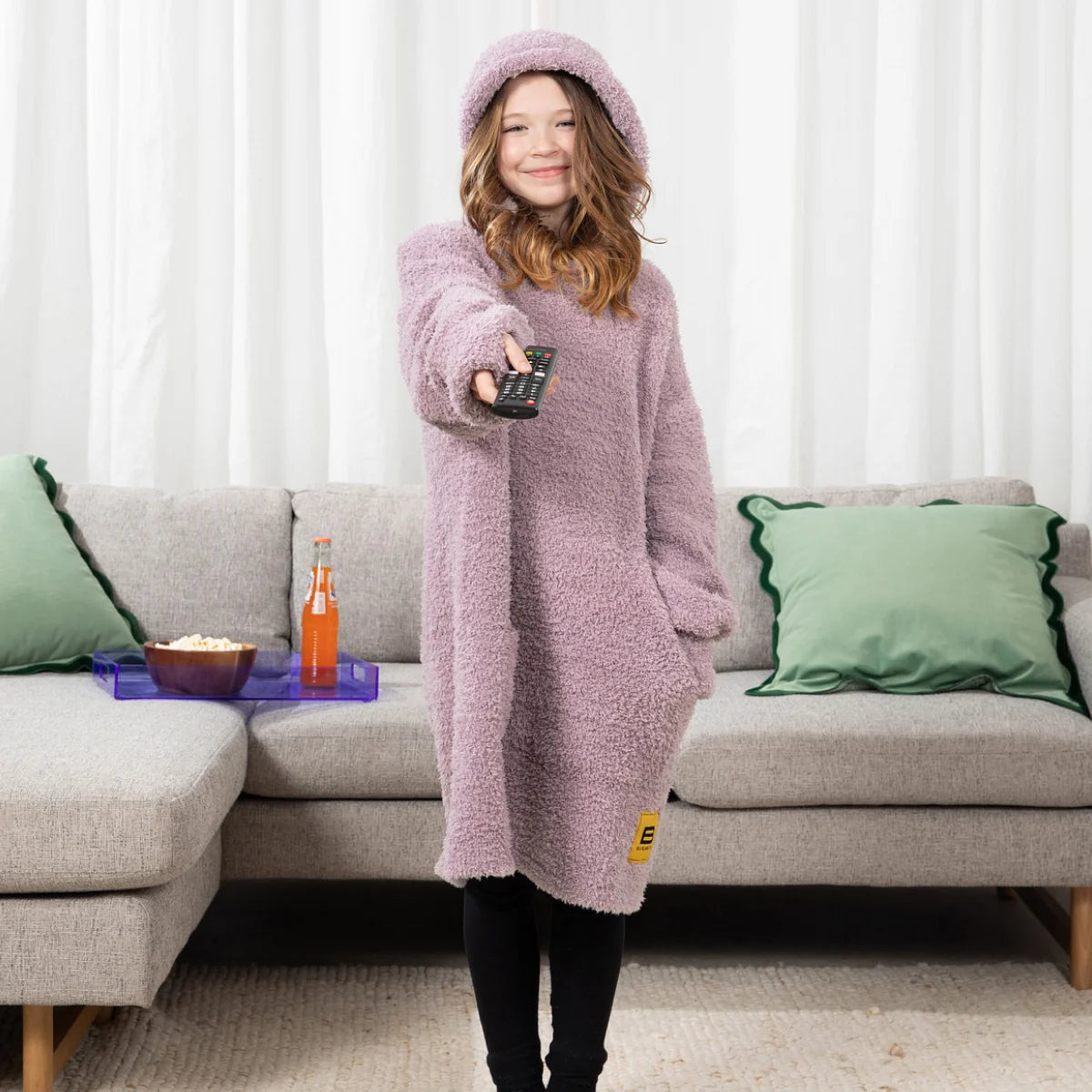 Child wearing mauve Premier Plush Hoodie with hood up, holding mug on bed, oversized blanket coverage