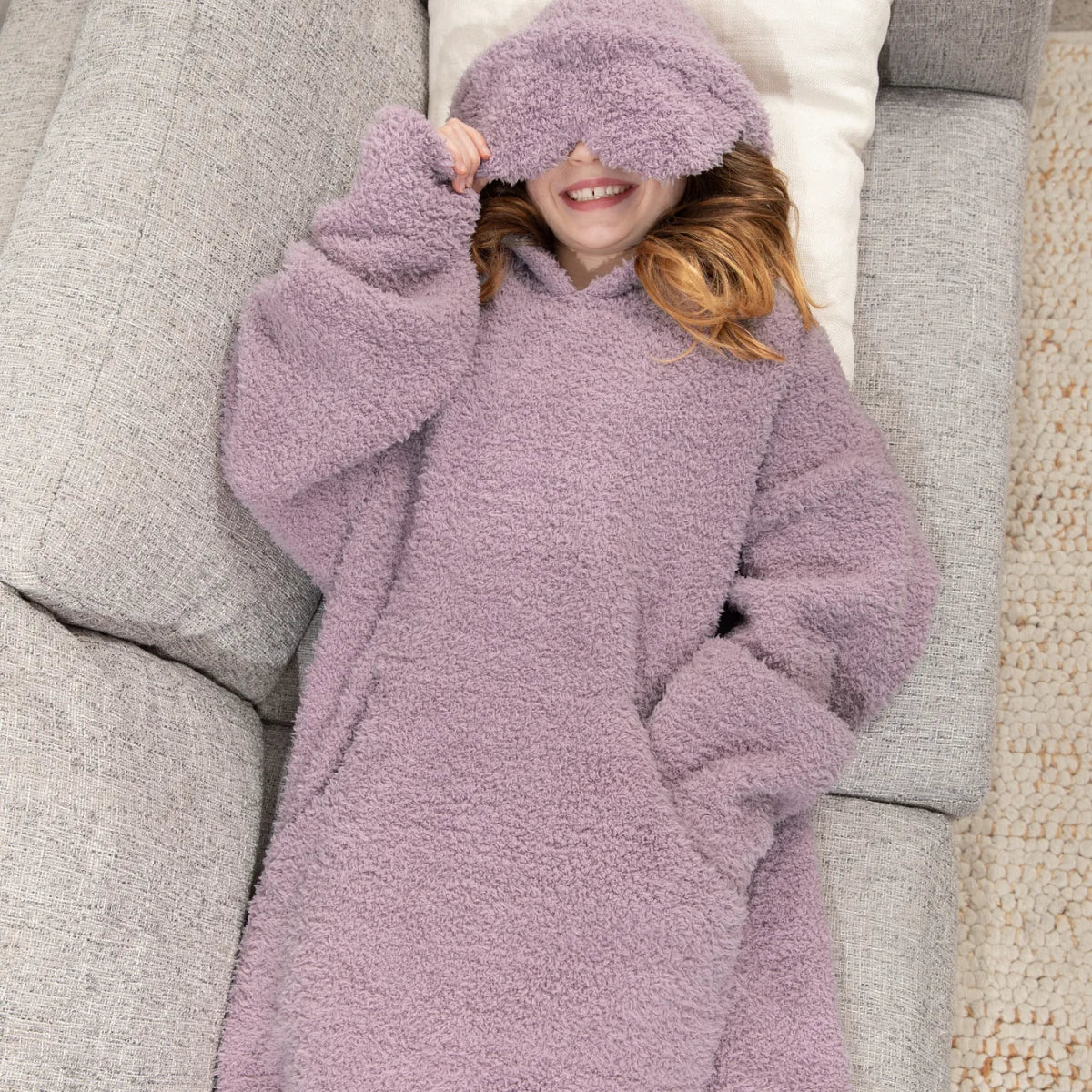 Child wrapped in soft mauve oversized hoodie blanket on white bedding, face covered by plush fabric hood