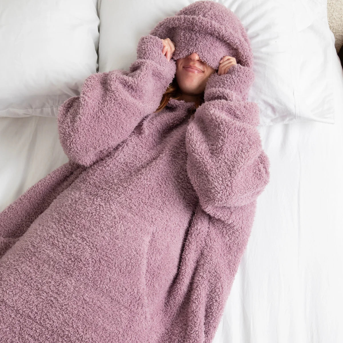 Person wrapped in mauve-pink oversized hoodie blanket on white bedding, showing plush texture and hood coverage