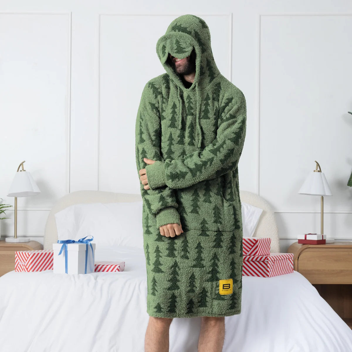 Person wearing sage green oversized blanket hoodie with forest pattern and hood pulled over head, standing on bed in bedroom setting