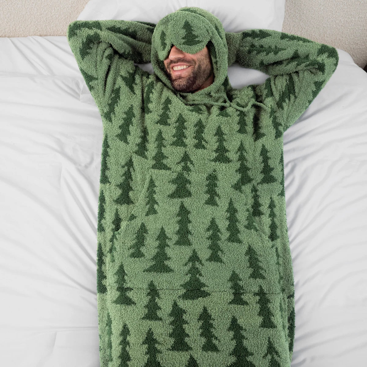 Man wrapped in sage green oversized hoodie blanket with forest tree pattern, lying on bed with hood covering eyes