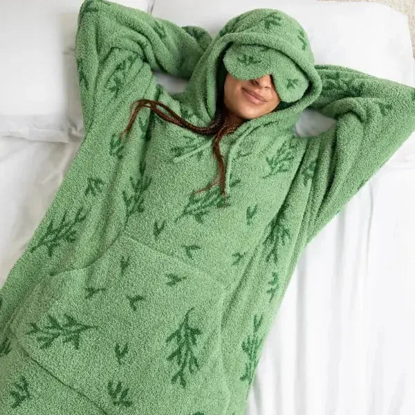 Child wrapped in sage green oversized blanket hoodie with fern pattern, hood pulled up, lying on white bedding