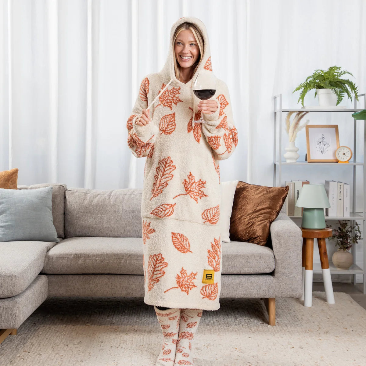 Woman wearing cream Premier Plush hoodie blanket with rust leaf pattern, holding wine glass on living room sofa