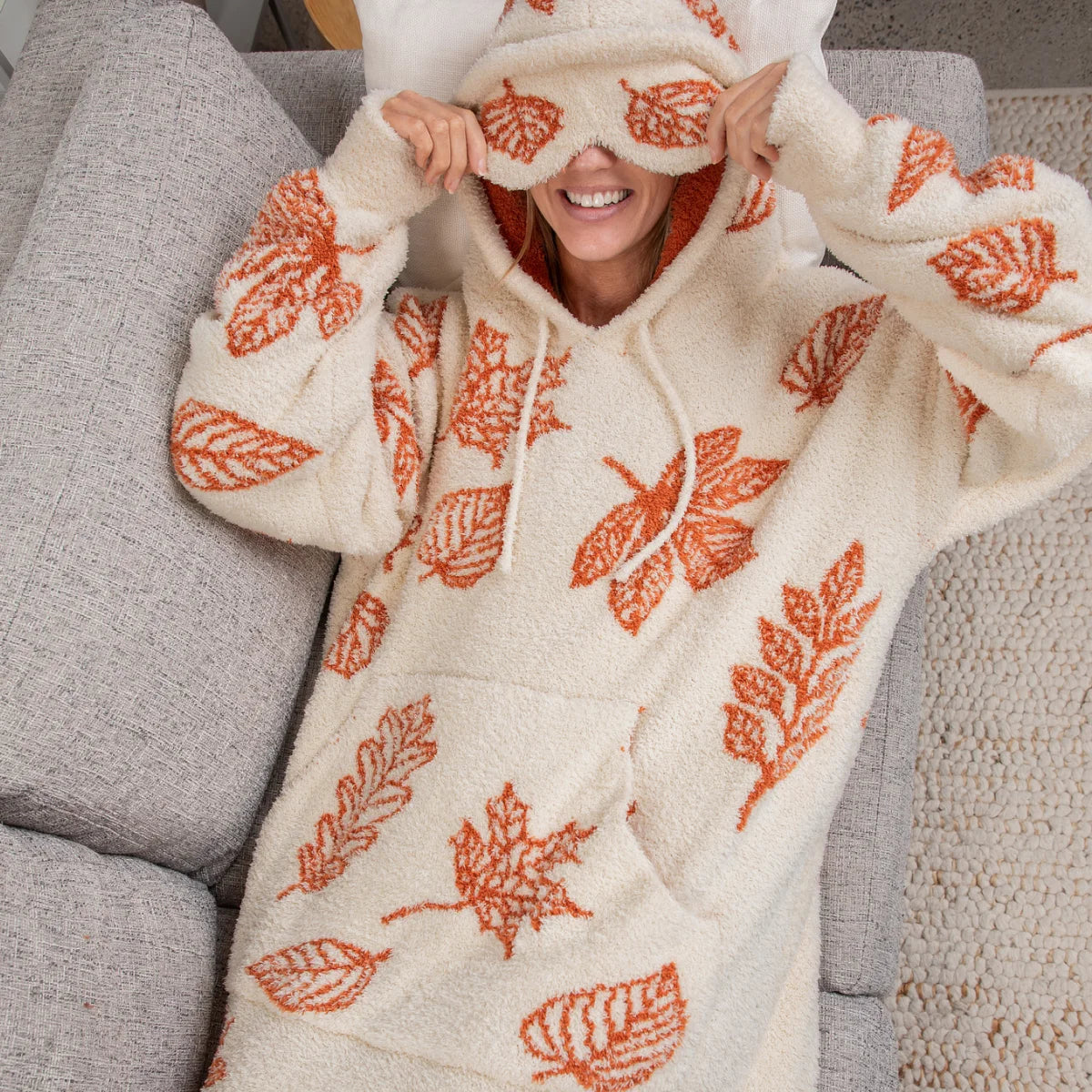 Woman wearing cream oversized hoodie blanket with orange leaf pattern, sitting on gray couch with hood pulled up