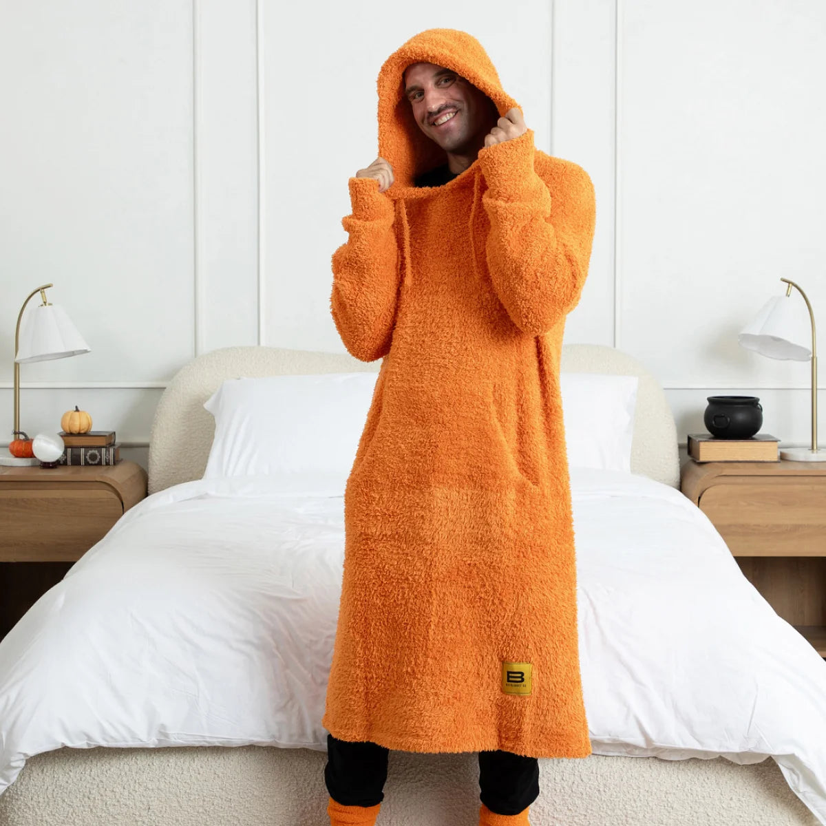 Person wearing orange oversized hoodie blanket with hood up, standing on bed with white pillows