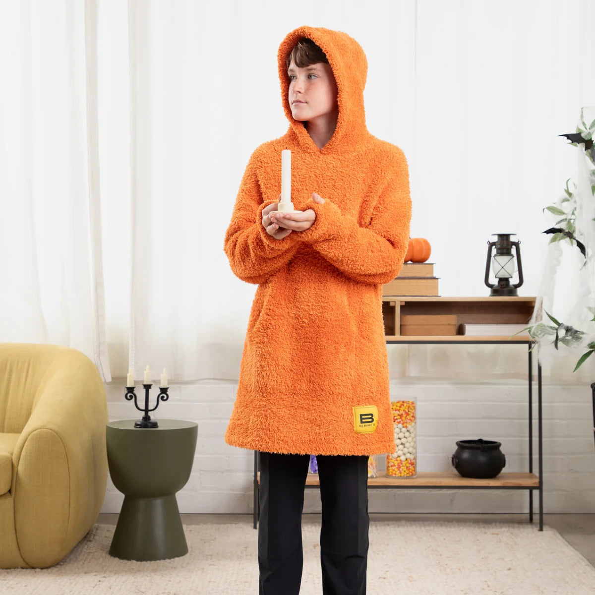 Person wearing oversized orange Premier Plush Hoodie Blanket with hood pulled up, standing on bed with white bedding