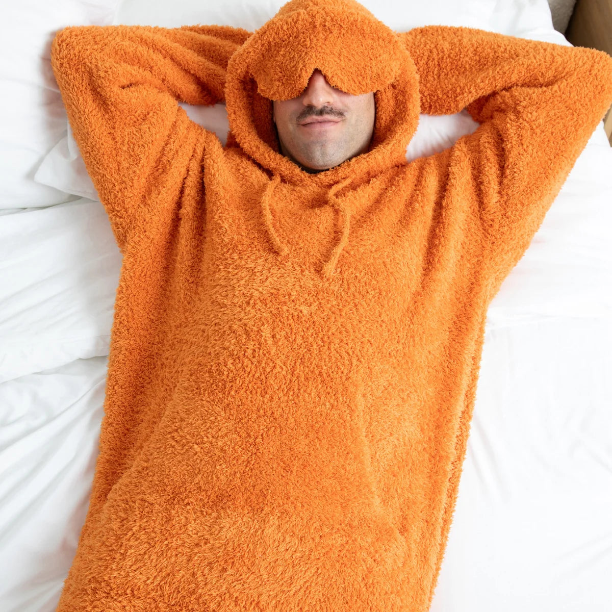 Person wrapped in oversized orange fuzzy hoodie blanket on white bedding, covering face with hood
