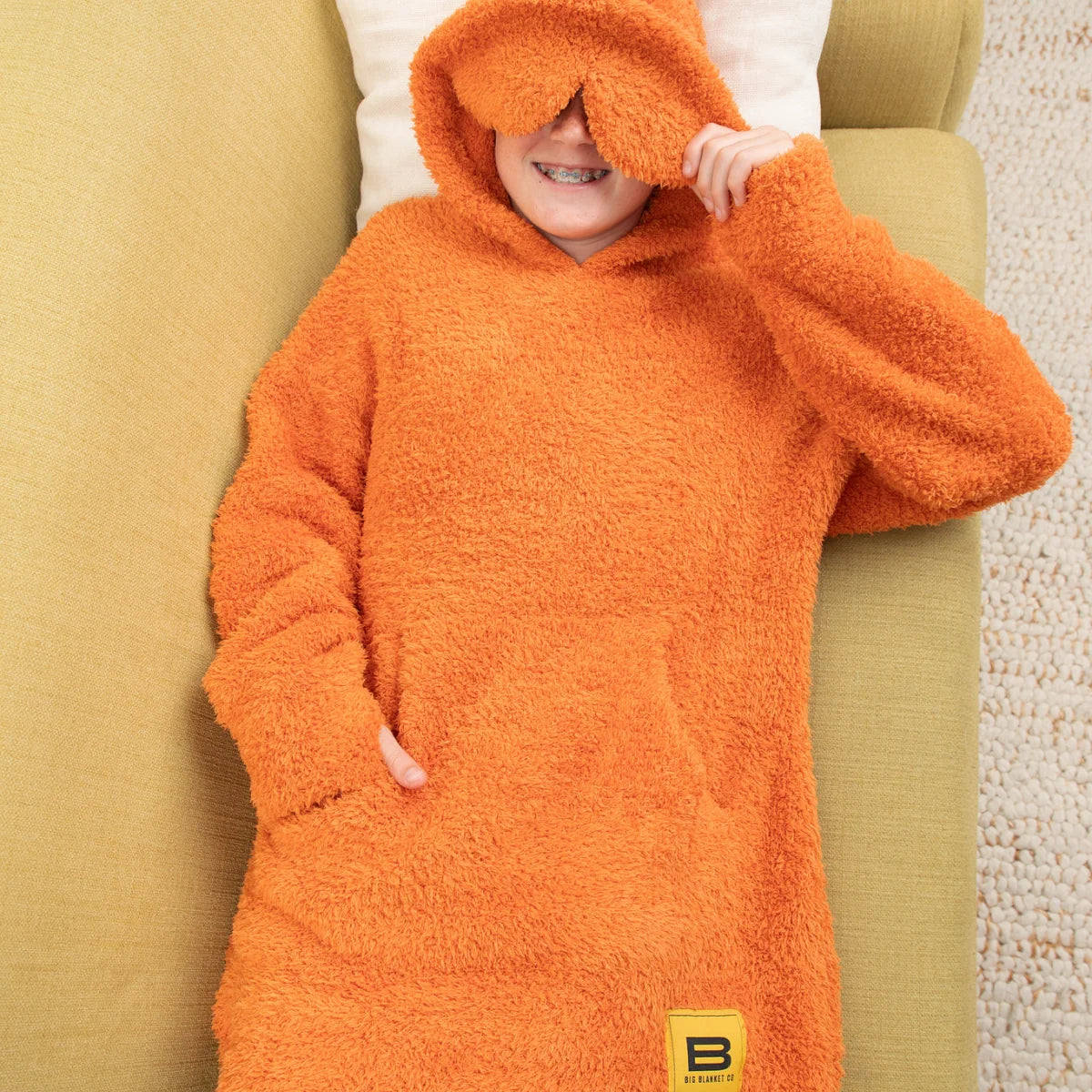 Person wrapped in oversized burnt orange fuzzy blanket with hood covering eyes, arms spread wide on white bedding
