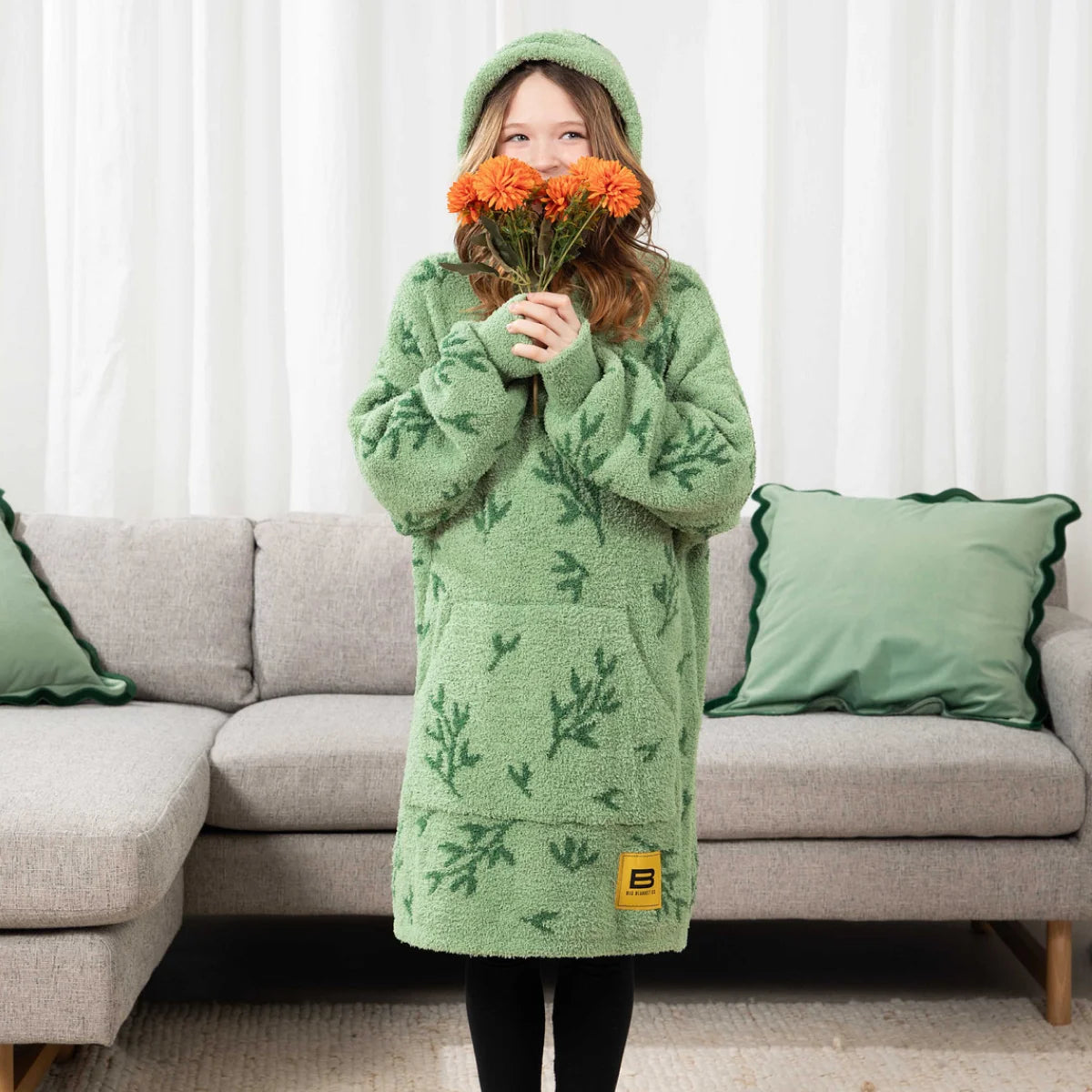 Woman wearing green oversized hooded blanket with leaf pattern and matching socks, standing in front of bed with white sheets