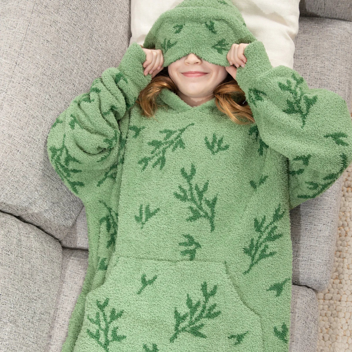 Person wrapped in sage green oversized hoodie blanket with fern leaf pattern and brown drawstring, head covered on white bedding