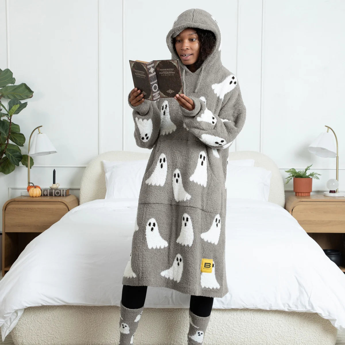 Gray oversized hoodie blanket with white ghost pattern, worn by person sitting on bed reading a book