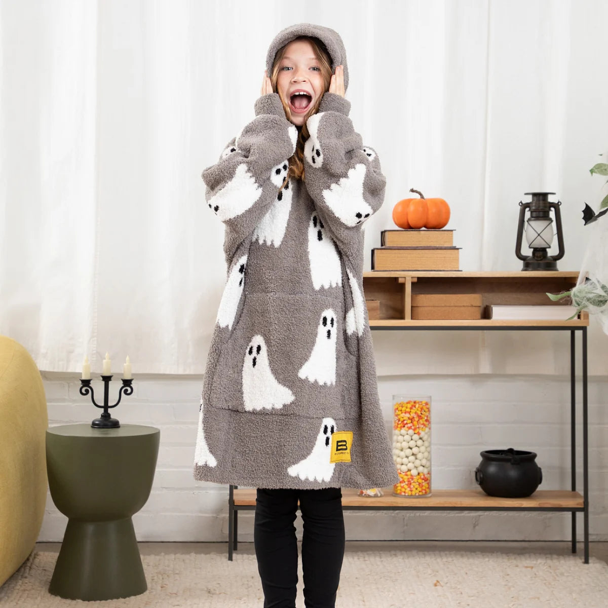 Person wearing gray Premier Plush hoodie blanket with white ghost pattern and hood, reading book on bed