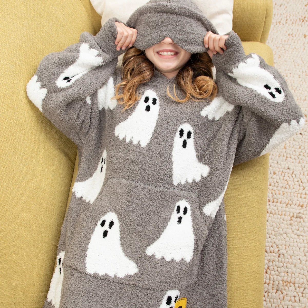 Child wrapped in gray oversized blanket with white ghost and black dot pattern, wearing attached hood, smiling on white bedding
