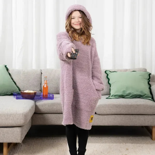 Woman wearing lavender Premier Plush Hoodie blanket with hood, holding remote control on beige sectional sofa