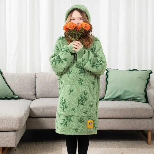 Girl wearing sage green oversized hoodie blanket with fern pattern, standing in front of sofa holding orange flowers