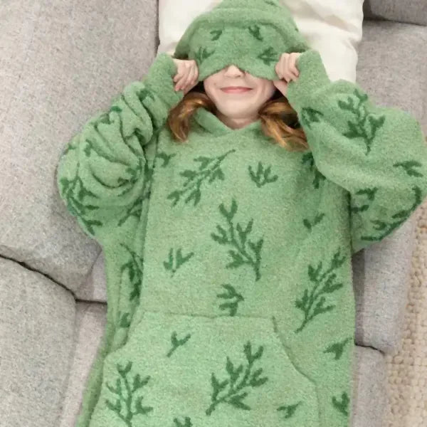 Child wearing sage green oversized hoodie blanket with fern leaf print, lying on gray couch with hood covering eyes
