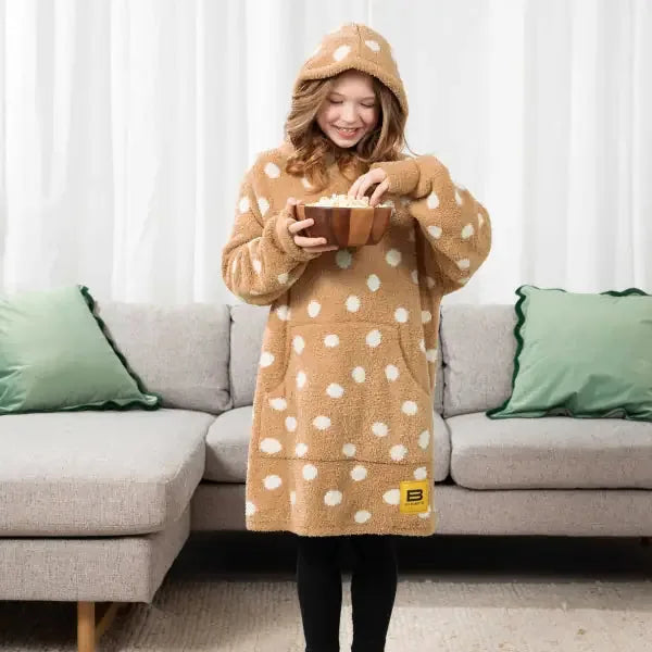 Child wearing tan oversized hoodie blanket with white polka dots, holding mug on couch