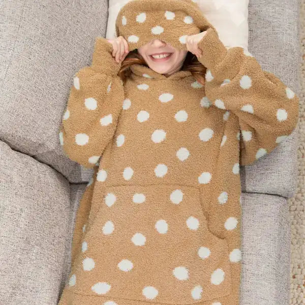 Child wearing oversized tan hoodie blanket with white polka dots, relaxing on beige couch