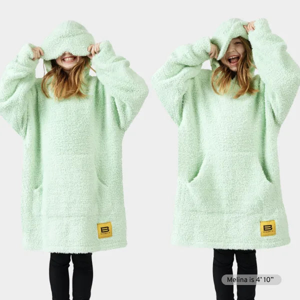 Mint green oversized hoodie blanket with hood and front pockets, modeled by person pulling hood over face and smiling