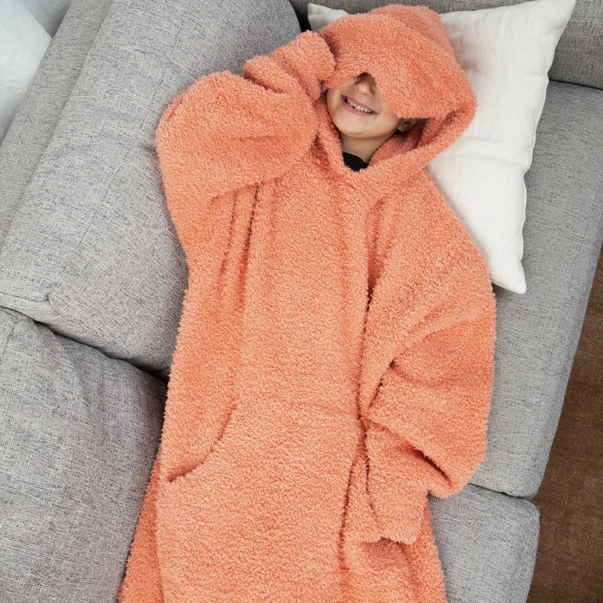Person wearing coral oversized hoodie blanket with front pouch and hood on bed