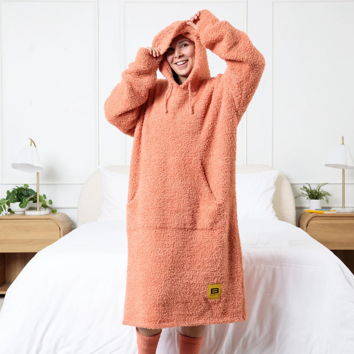 Person wearing coral oversized hoodie blanket with cozy texture and large hood, standing on bed