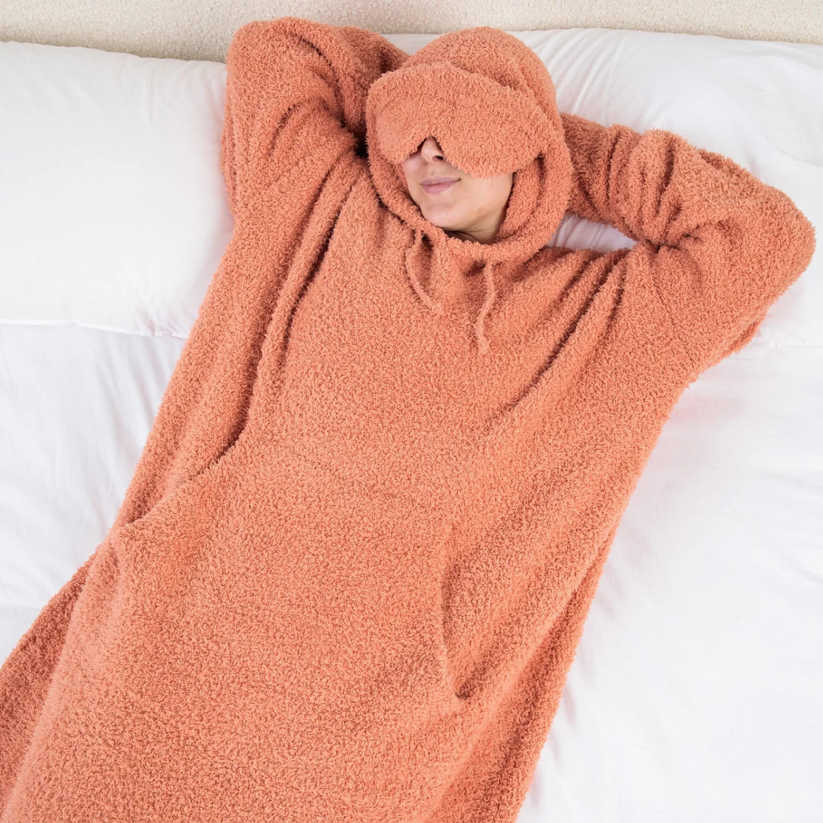 Person wrapped in oversized melon-colored plush hoodie blanket on white bedding, face covered by hood