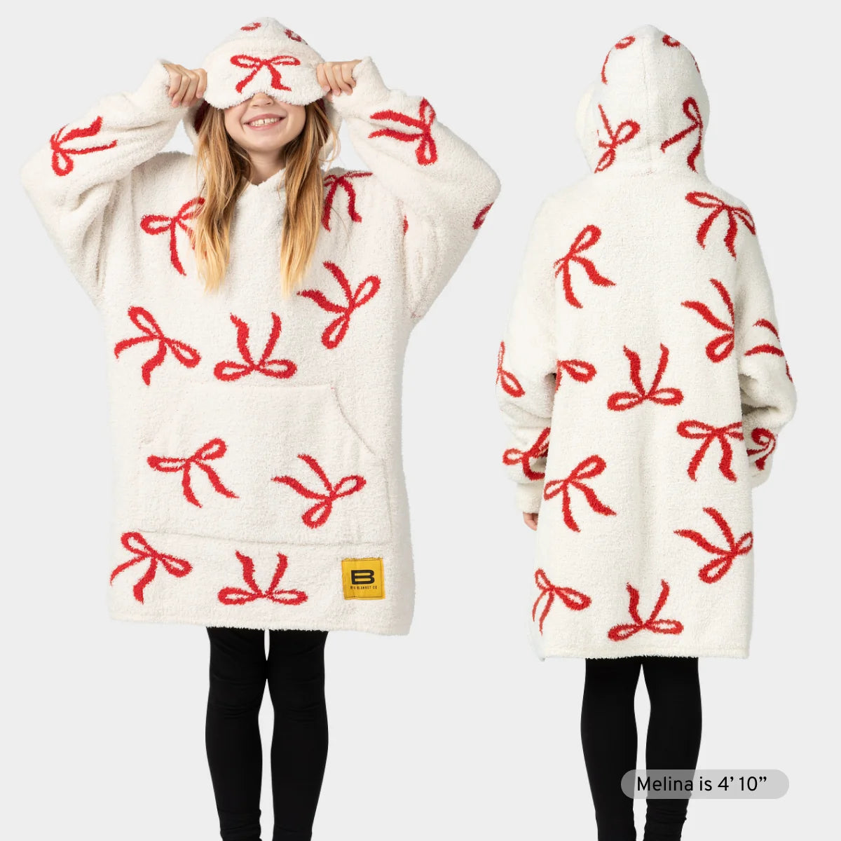 Two people wearing cream oversized hoodie blankets with red bow pattern, matching socks, and hoods pulled up