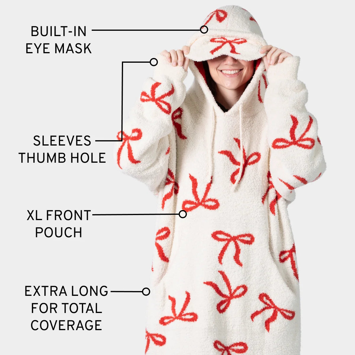 Cream oversized hoodie blanket with red bow pattern, featuring built-in eye mask, thumb holes, XL front pouch, and extra-long coverage