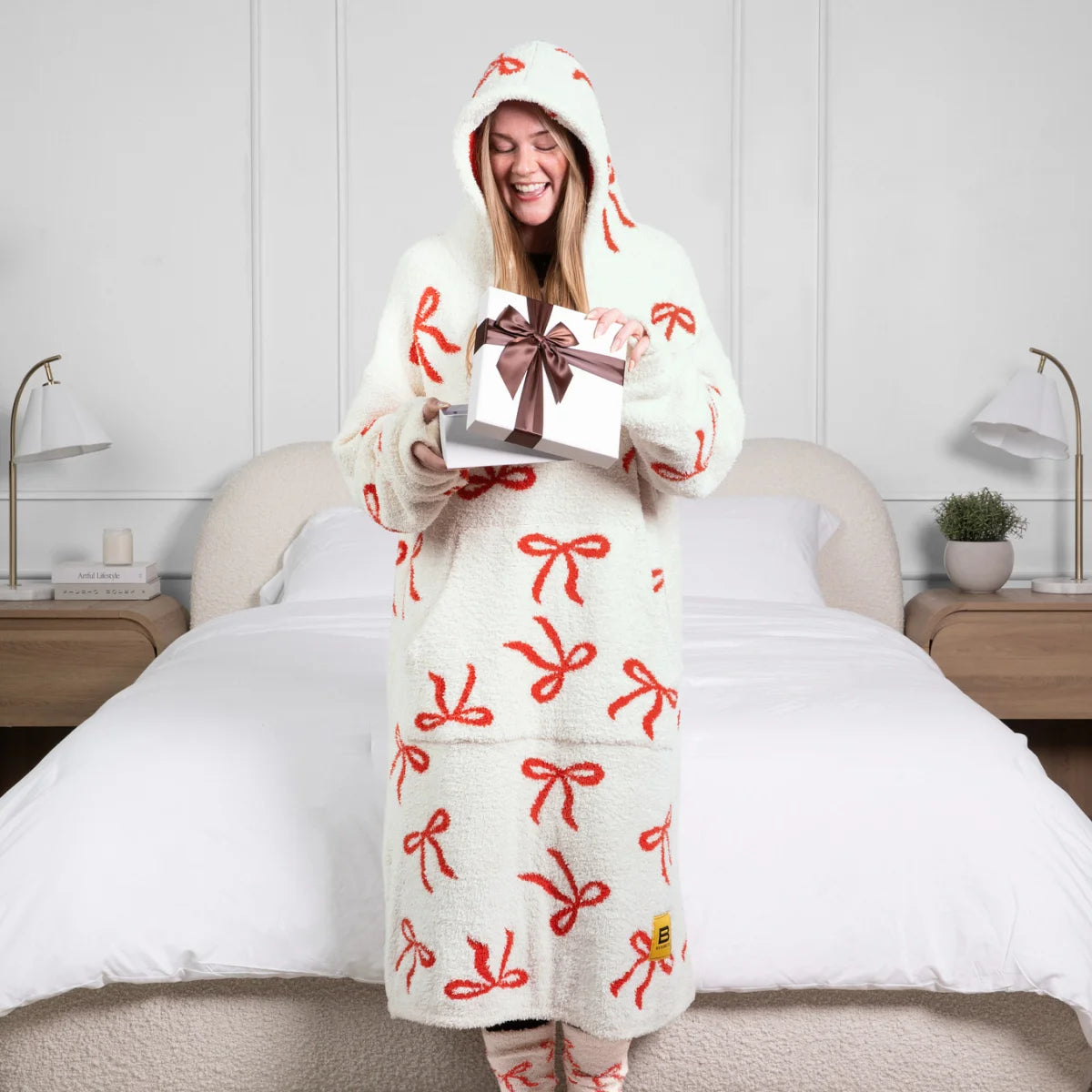 Woman wearing cream oversized hoodie blanket with red bow pattern, holding gift on bed