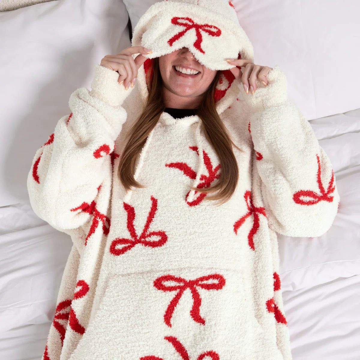 Cream oversized hoodie blanket with red bow pattern, woman wearing it with hood up covering eyes, cozy plush fabric