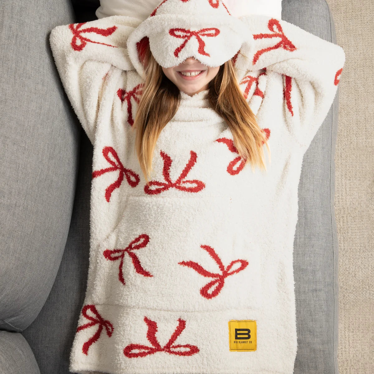 Woman wearing cream oversized hoodie blanket with red bow pattern and matching hood, smiling on white bedding