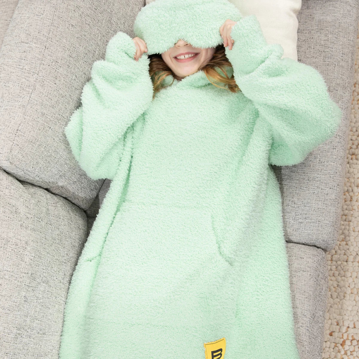 Child wrapped in soft mint green oversized hoodie blanket with hood covering eyes, lying on gray couch