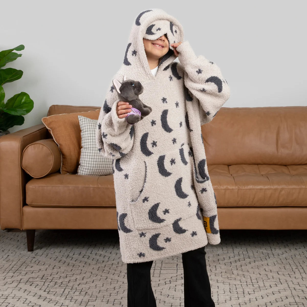 Child wearing cream oversized hoodie blanket with moon and star pattern, standing in living room holding stuffed animal