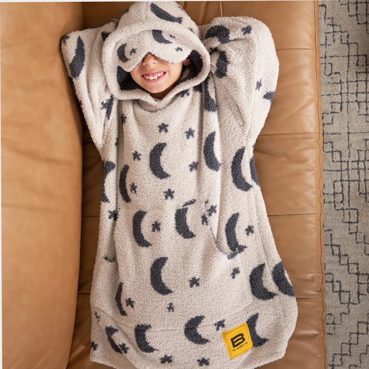 Child wrapped in cream hooded blanket with moon and star pattern, sitting on tan leather couch