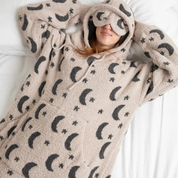 Person wrapped in beige oversized hoodie blanket with moon and star print, hood covering head